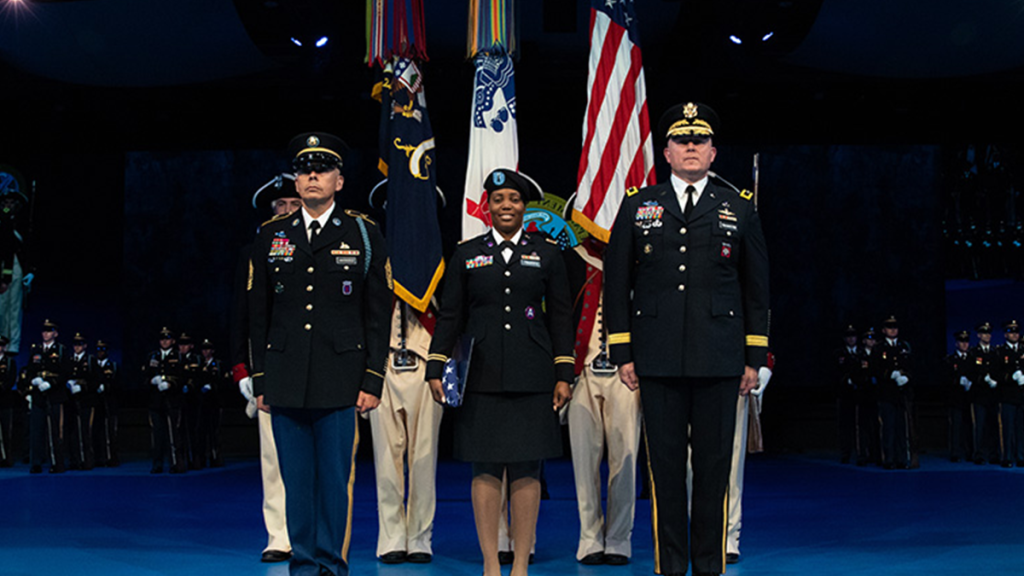 Sheree Stewart on stage standing next to two distinguished military individuals in full regalia.