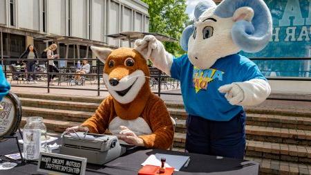 A ram mascot, Rameses Jr., holding the ear of another mascot, Poetry Fox, at Arts Everywhere Day.