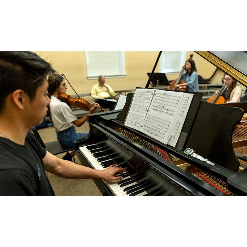 Students in a music class playing a variety of instruments. In the foreground is a student playing the piano and reading sheet music.